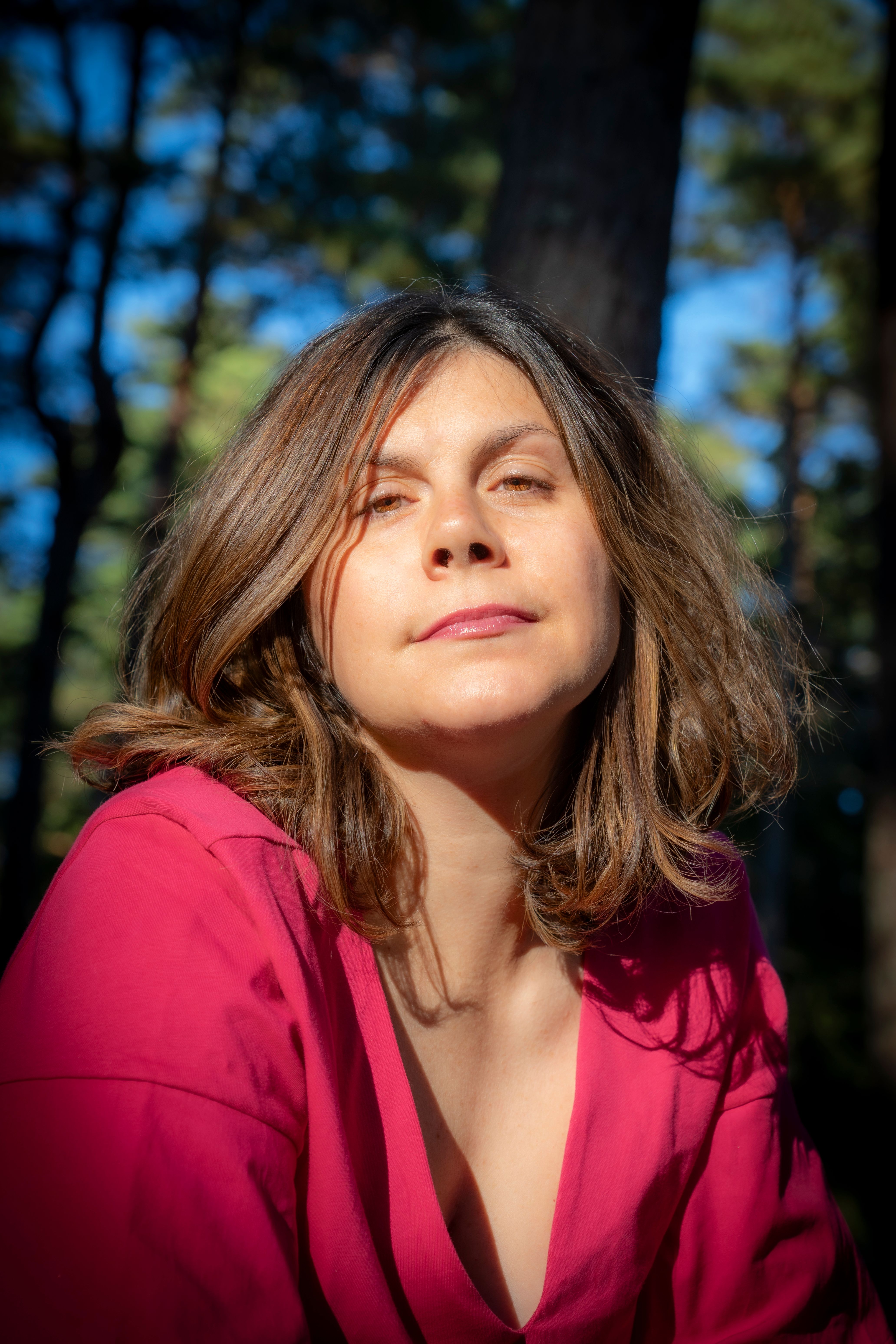 A woman in a pink shirt poses outdoors, surrounded by a lush green forest and bright sunlight.