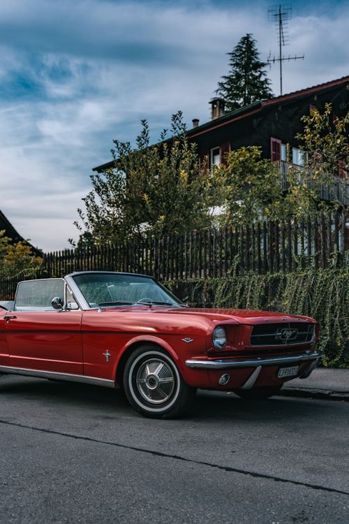 Free Red Ford Mustang cabriolet parked on a street with vintage charm and lush greenery. Stock Photo