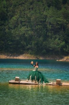 A woman in a green gown stands on a wooden dock by a serene lake, surrounded by lush forest.