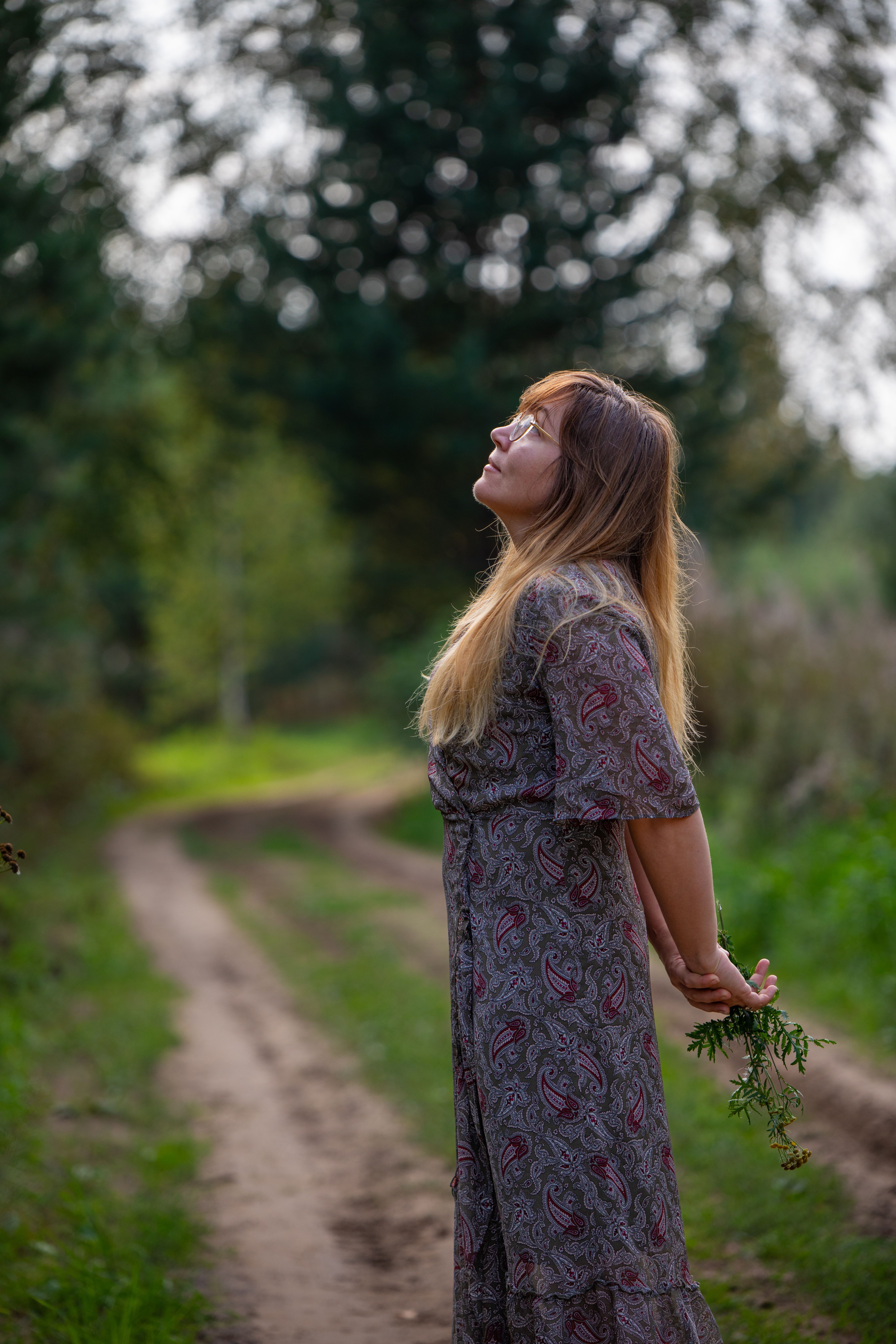 Peaceful portrait of a woman in a sundress standing on a rural dirt road, surrounded by nature.