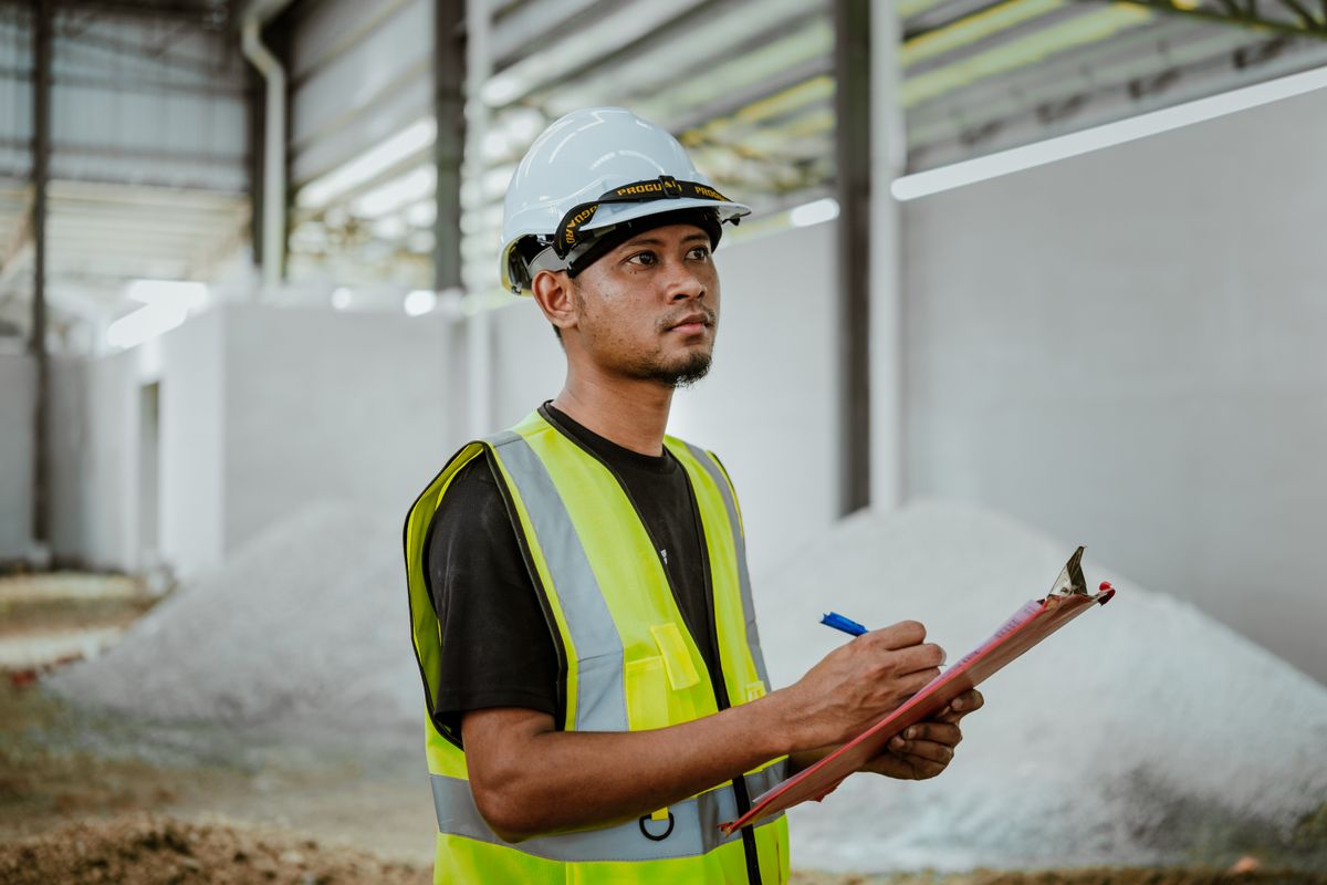 Malaysian site supervisor in hi-vis and hard hat checking a clipboard during an inspection walk at an industrial site in Selangor