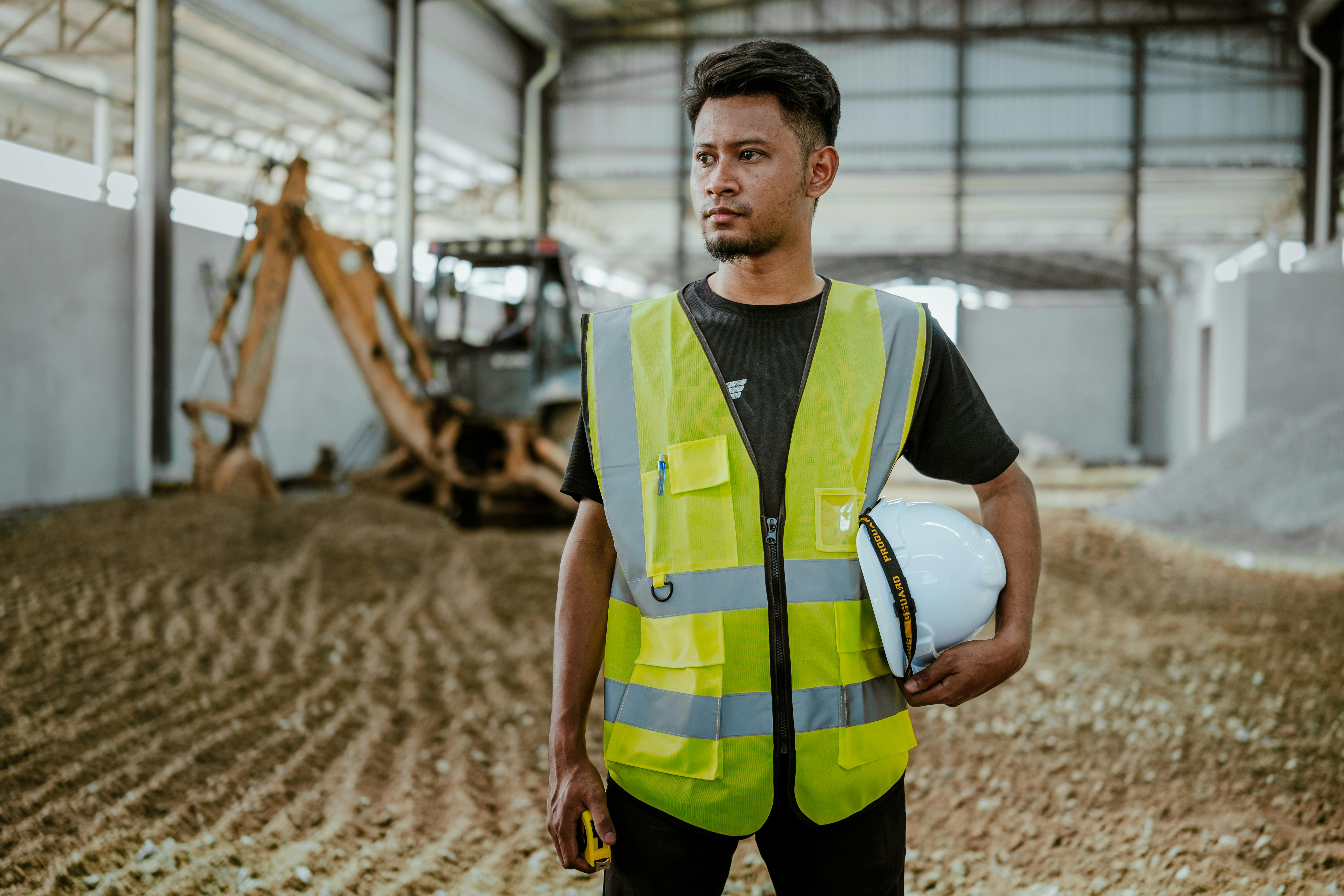 a worker at a construction site wearing a neon green vest and holding a safety hat in his arm