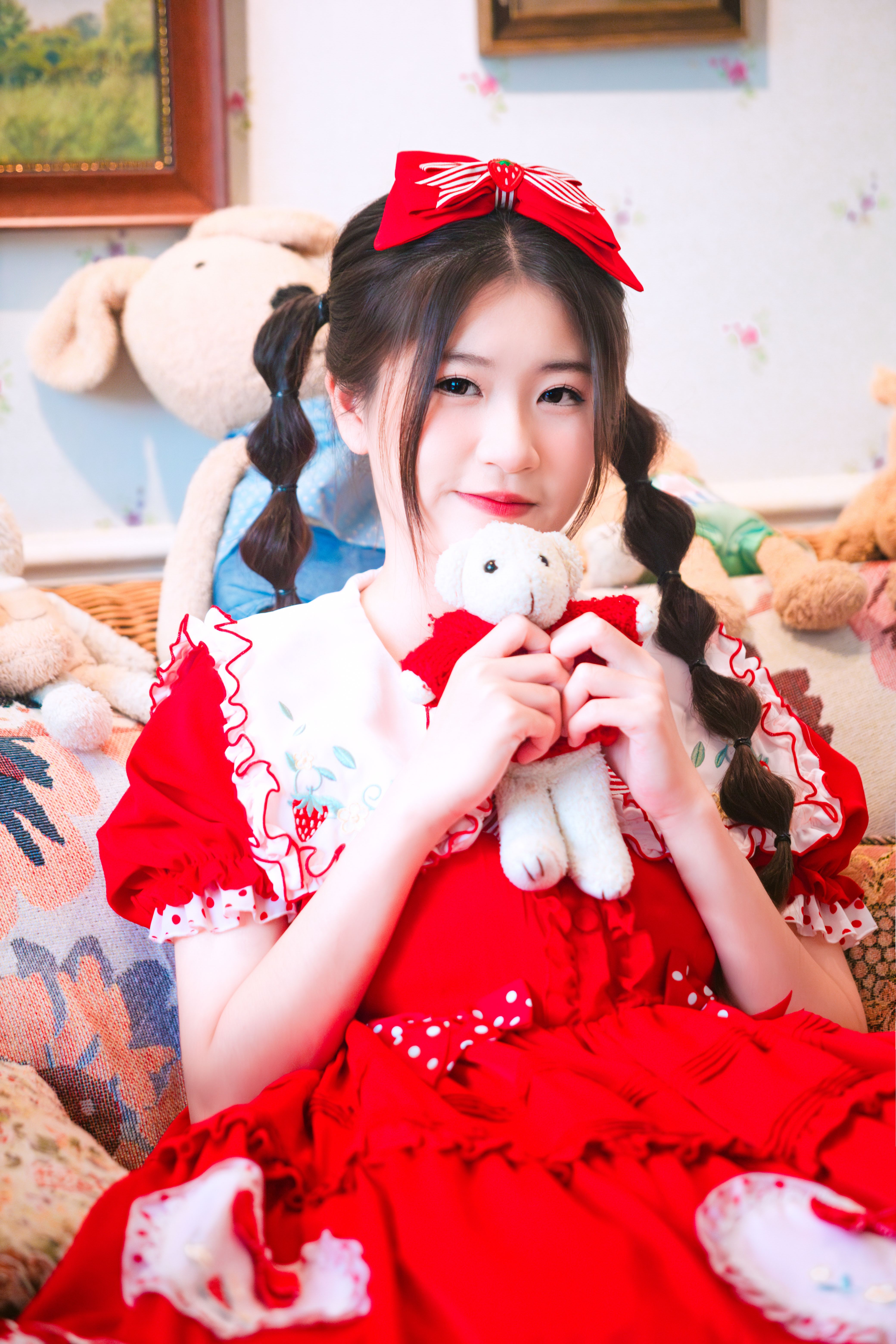 A cute girl in a red dress holding a teddy bear, surrounded by soft toys.