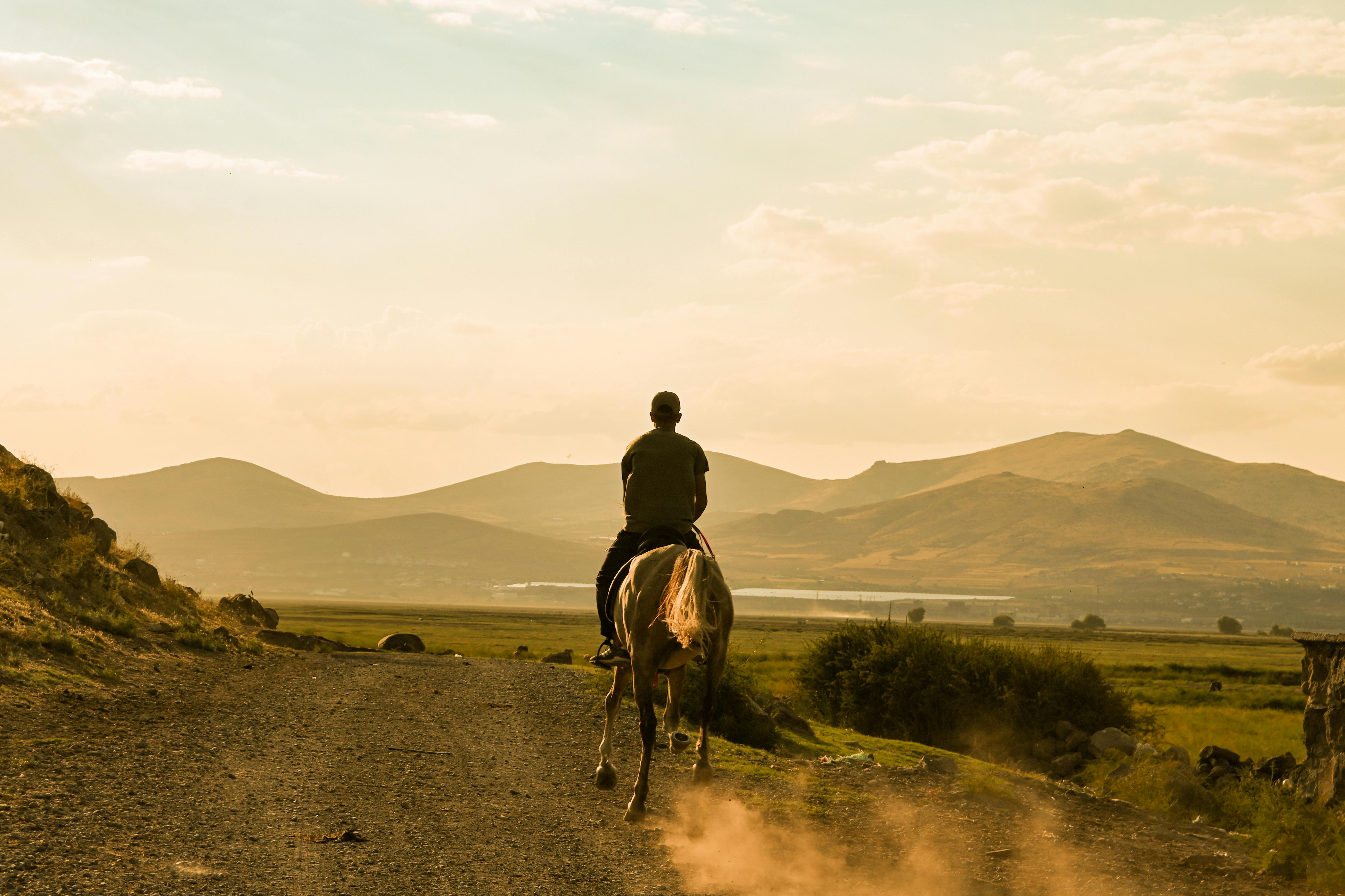 A man riding a horse on a dusty path with mountains in the background during sunset.