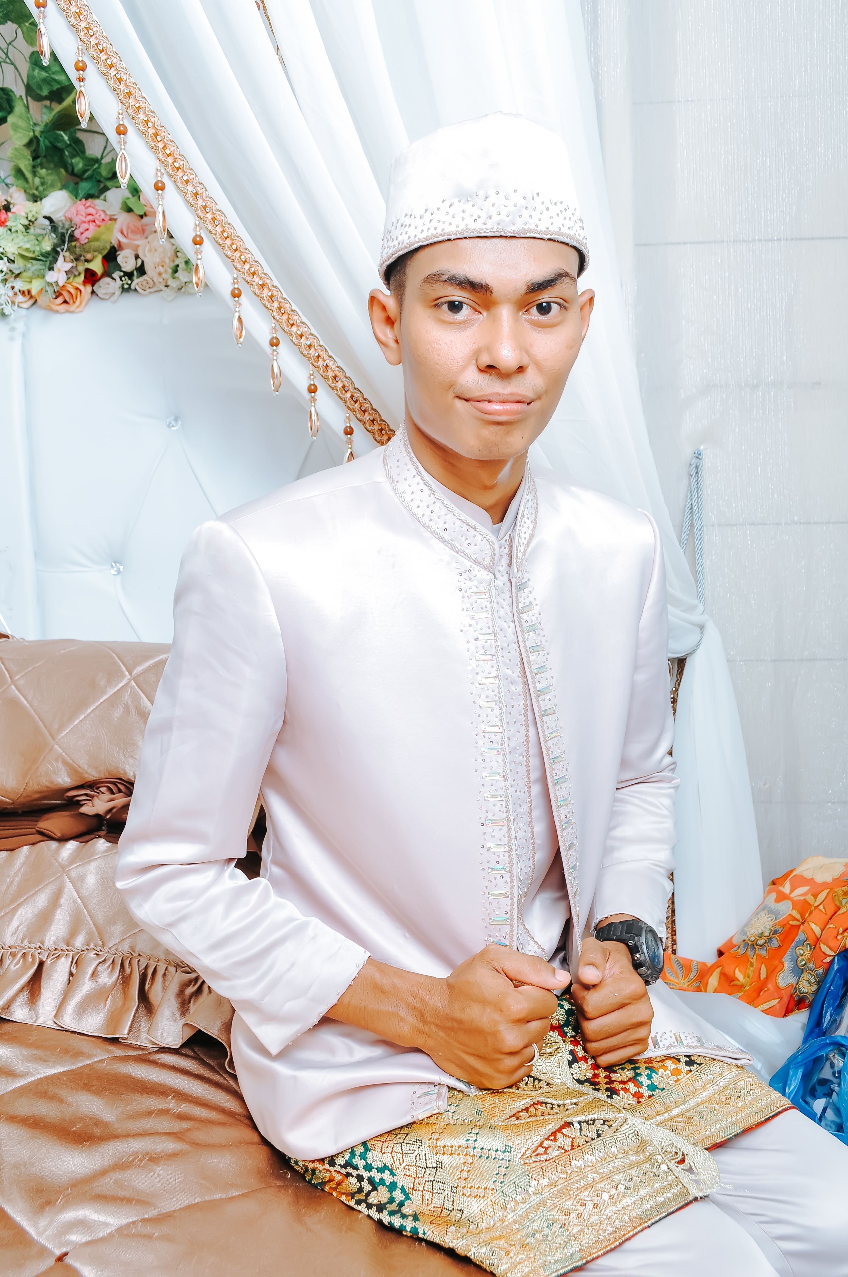 Young man in traditional attire seated, white background with floral decor.