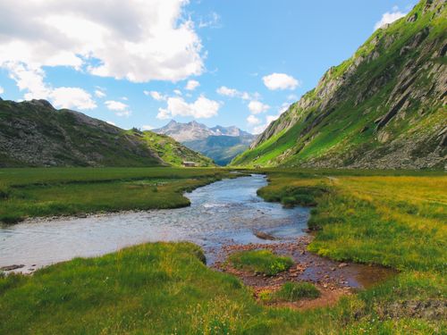 Free Tranquil river flowing through a lush green valley between majestic mountains under a clear blue sky. Stock Photo
