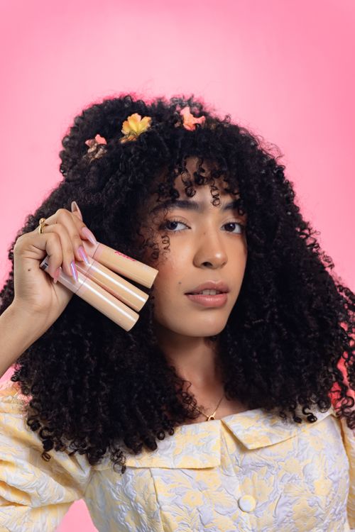 Free Portrait of a young woman with curly hair holding makeup items, set against a pink background. Stock Photo