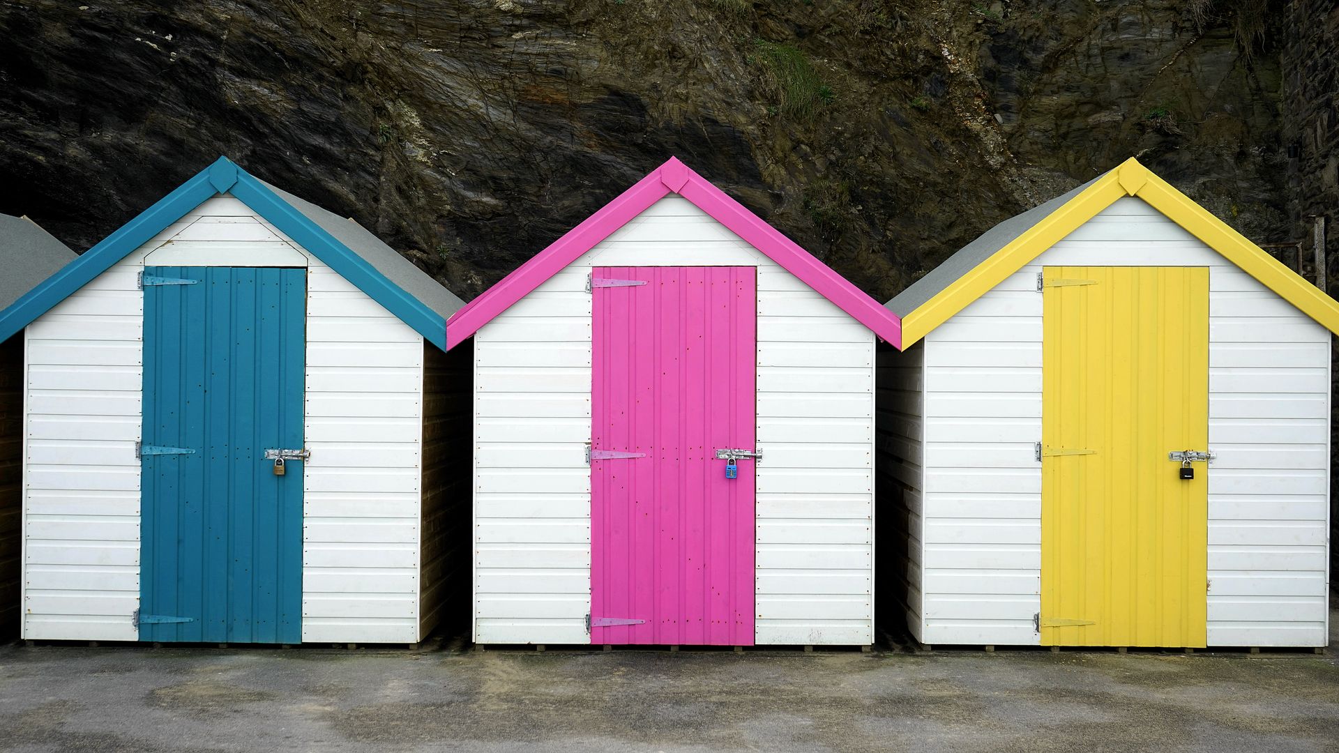 Three vibrant wooden beach huts with colorful doors in a row against a rocky backdrop.