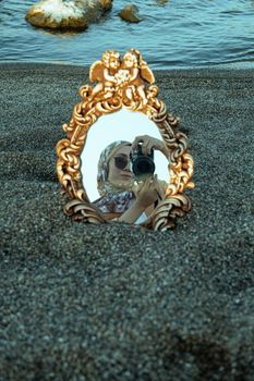 An ornate mirror reflecting a woman with a camera on a sandy beach, capturing a unique seaside moment.