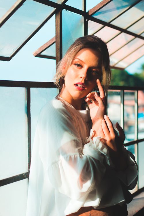 Free Artistic portrait of a woman posing by glass windows in a sunlit indoor setting, showcasing elegant fashion and style. Stock Photo