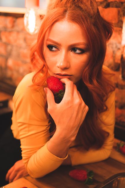 A redhead woman in a cozy cafe holding a strawberry, creating a warm and alluring scene.