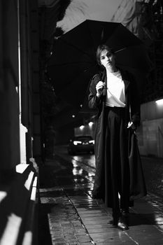 Black and white portrait of an elegant woman holding an umbrella on a dimly lit rainy street.