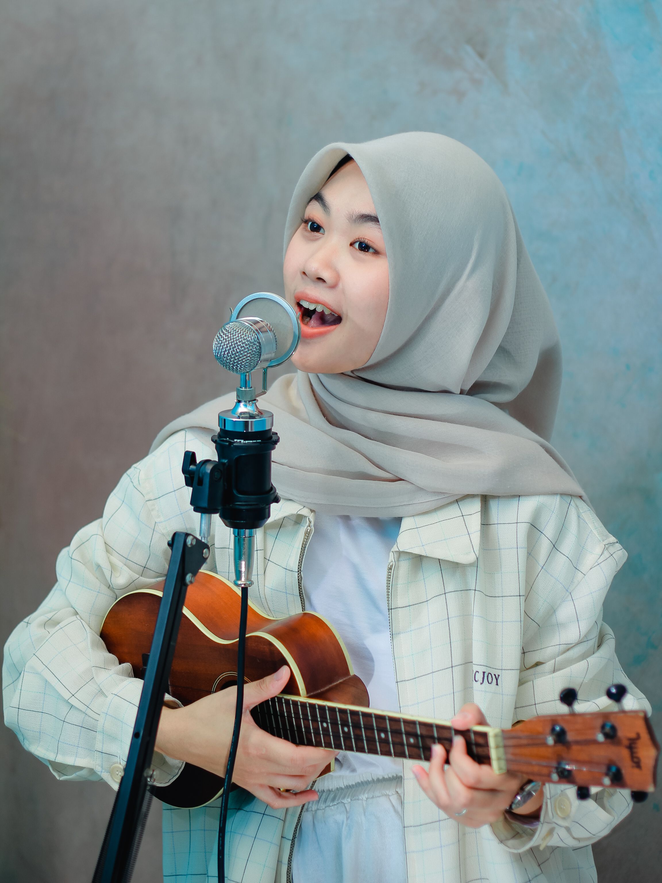 A young woman wearing a hijab plays the ukulele and sings into a microphone, showcasing musical talent.