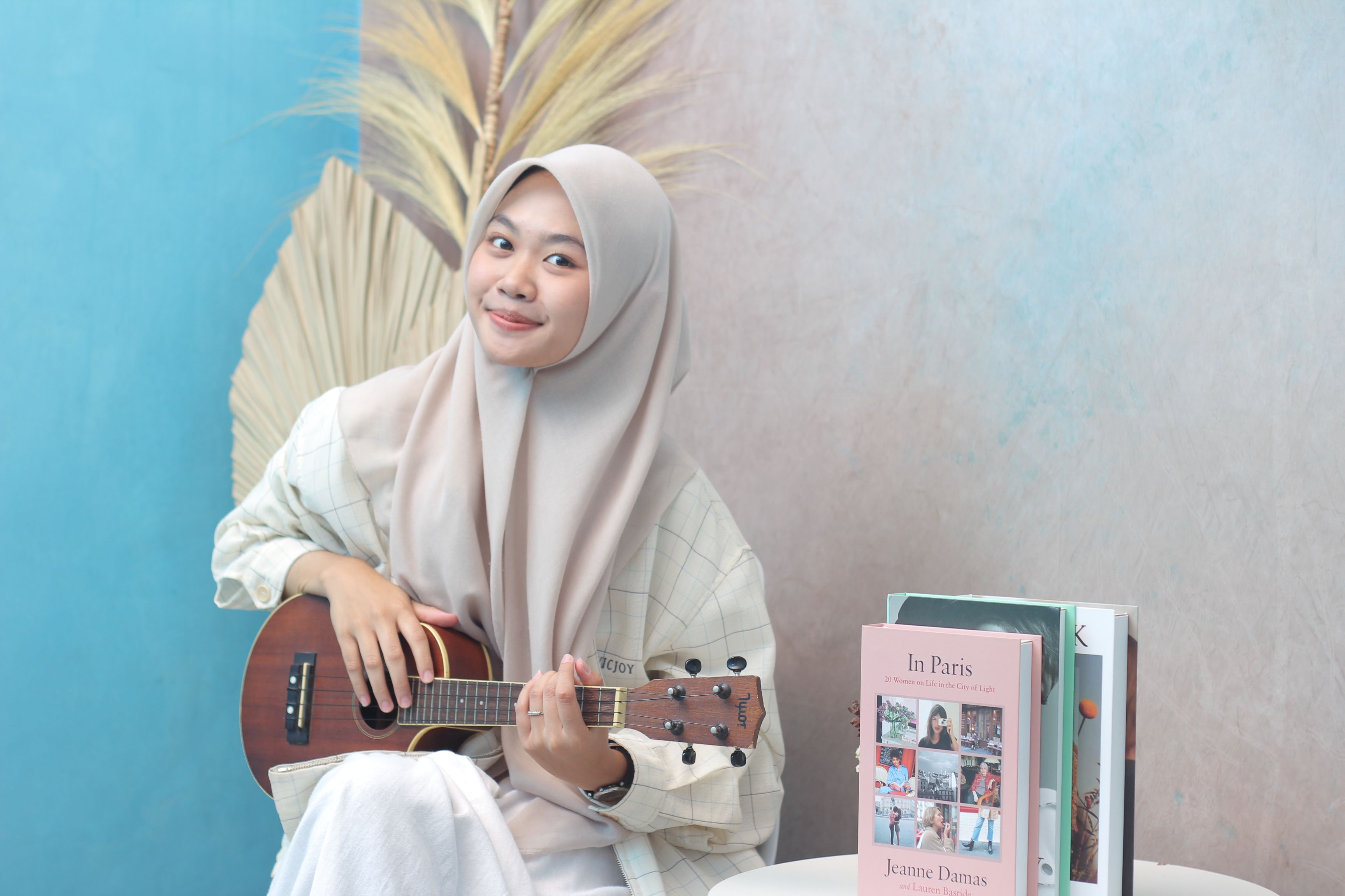 Smiling woman in hijab playing a ukulele with books and a creative background.