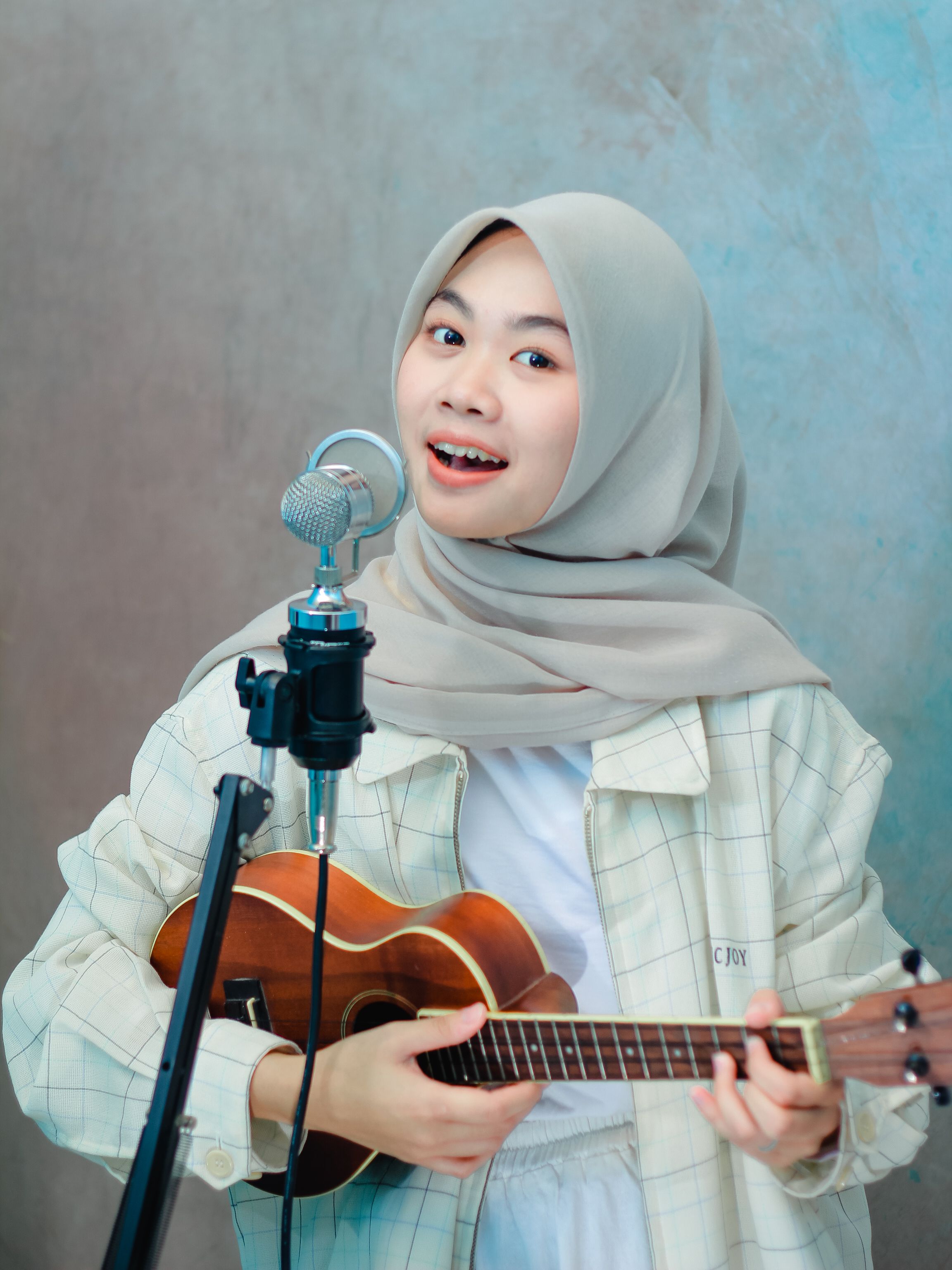Cheerful young woman wearing hijab, playing ukulele and singing into microphone.
