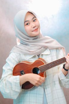 Charming young woman with a hijab playing a ukulele indoors, exuding a joyful and relaxed vibe.