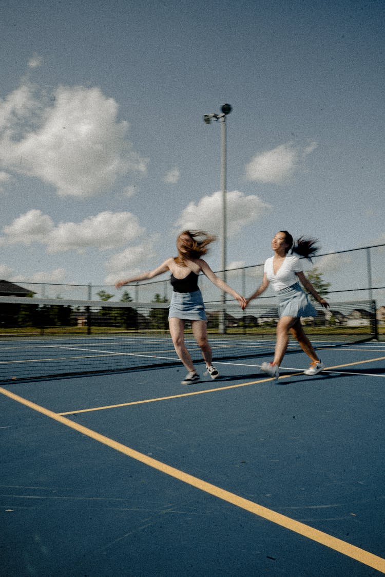 Woman Wearing Black String Strap Top On Tennis Court Besides Woman Wearing White Top