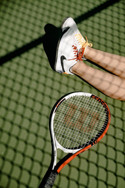 免费 人的脚穿白色和黑色的耐克低帮运动鞋，旁边缺少白色和橙色的威尔逊网球拍 素材图片