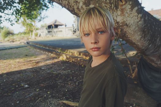 A blonde boy standing outdoors by a tree, capturing a quiet moment.