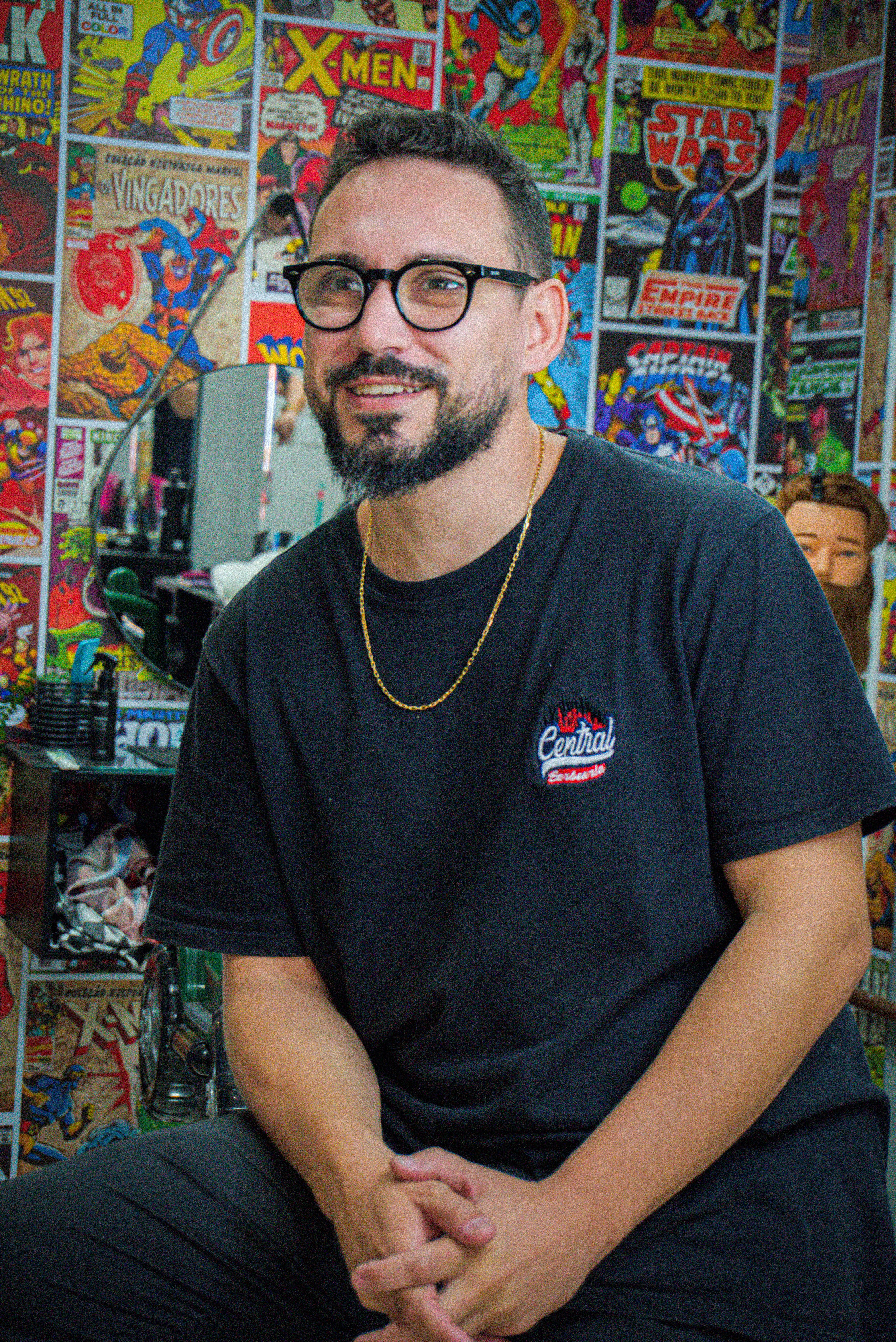 Man sitting in comics-themed barbershop, wearing glasses and casual attire, enjoying a relaxed moment.
