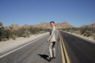 阳光明媚，一名身穿西装的男子走在荒无人烟的沙漠高速公路上，背景是山脉