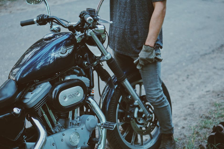 A person in casual attire standing next to a vintage motorcycle on an outdoor road.