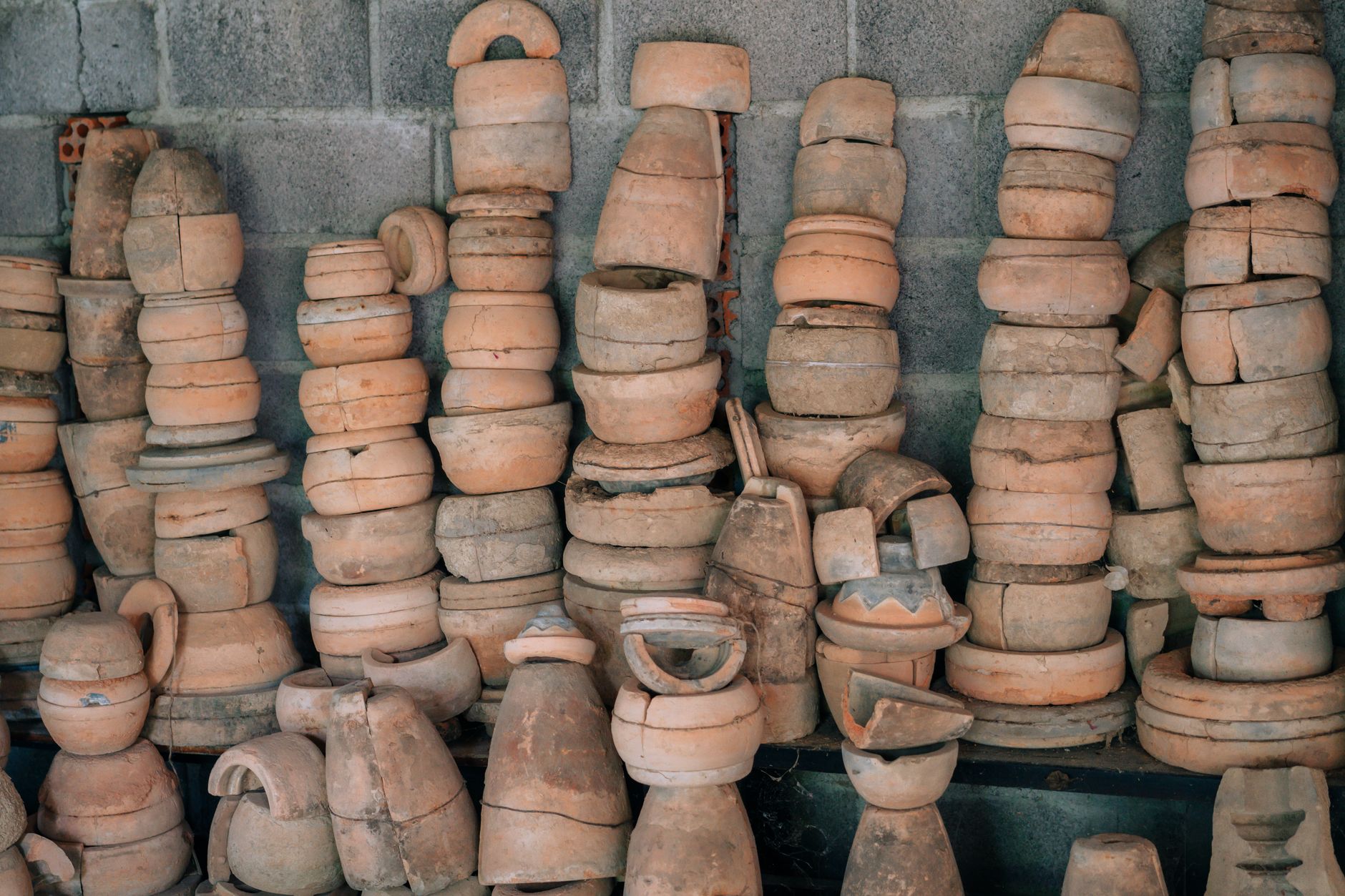 An array of handmade clay pottery pieces stacked against a cement wall, showcasing rustic craft.