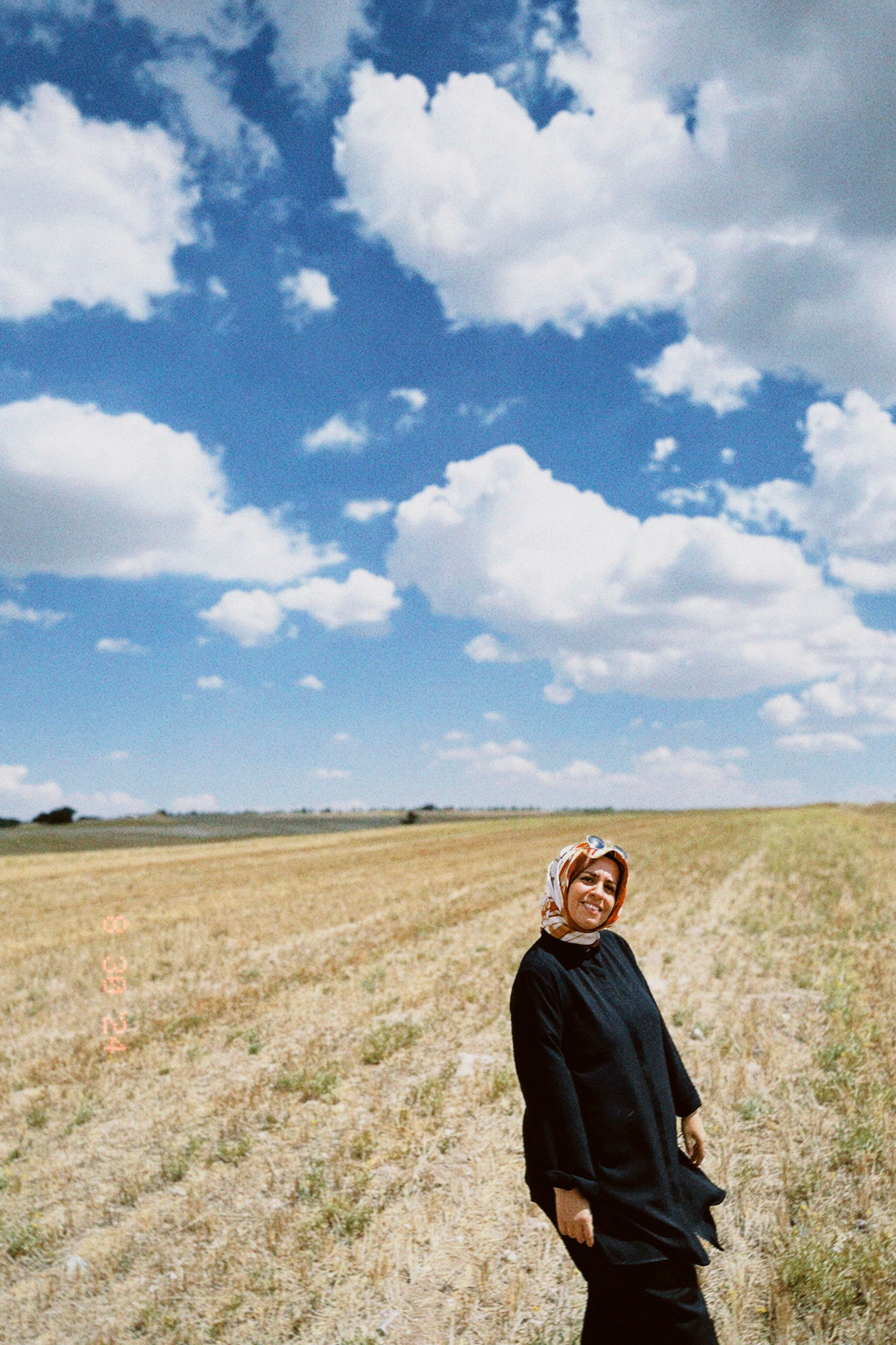 A young woman in a hijab smiles brightly in a sunlit countryside field under a blue sky.
