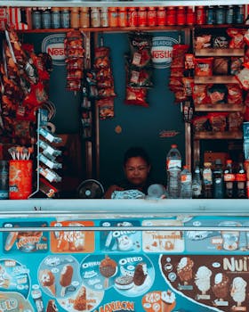Woman managing a vibrant snack shop in Barcelona displaying various chips and beverages.
