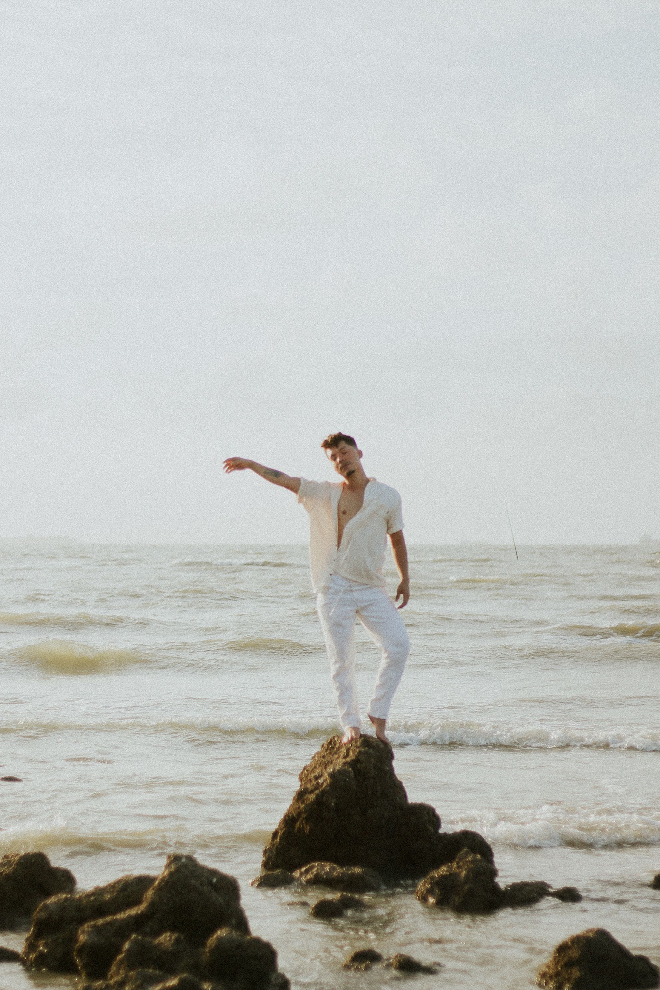 A man in summer attire stands on a rock by a serene ocean, exuding a relaxed vibe.