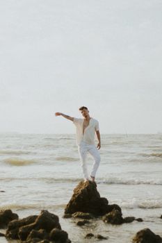 A man in summer attire stands on a rock by a serene ocean, exuding a relaxed vibe.