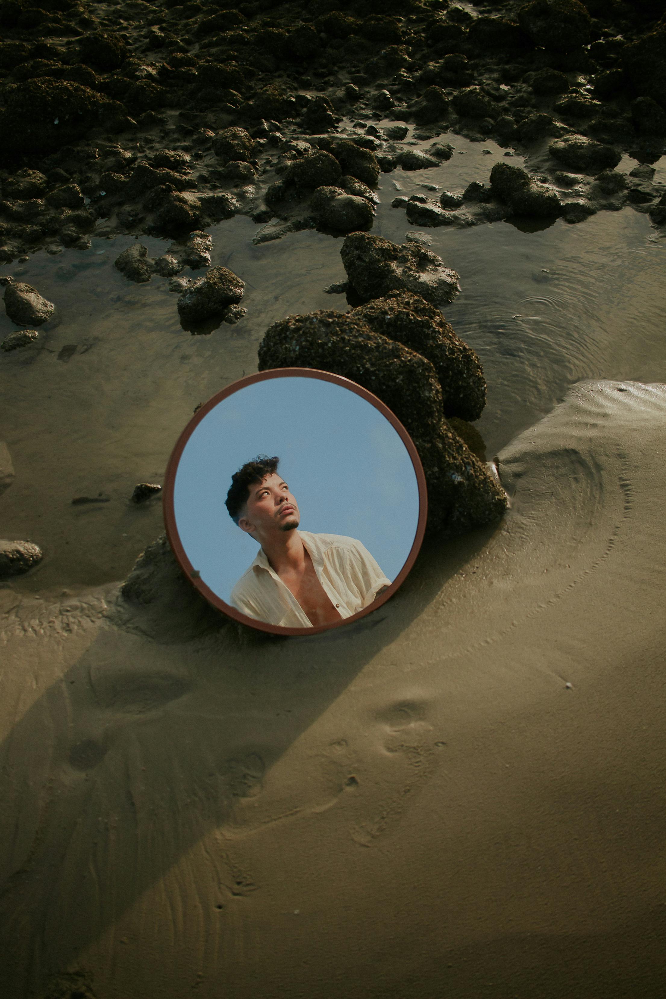 A creative portrait reflecting a man in a mirror on a sandy beach surrounded by rocks.