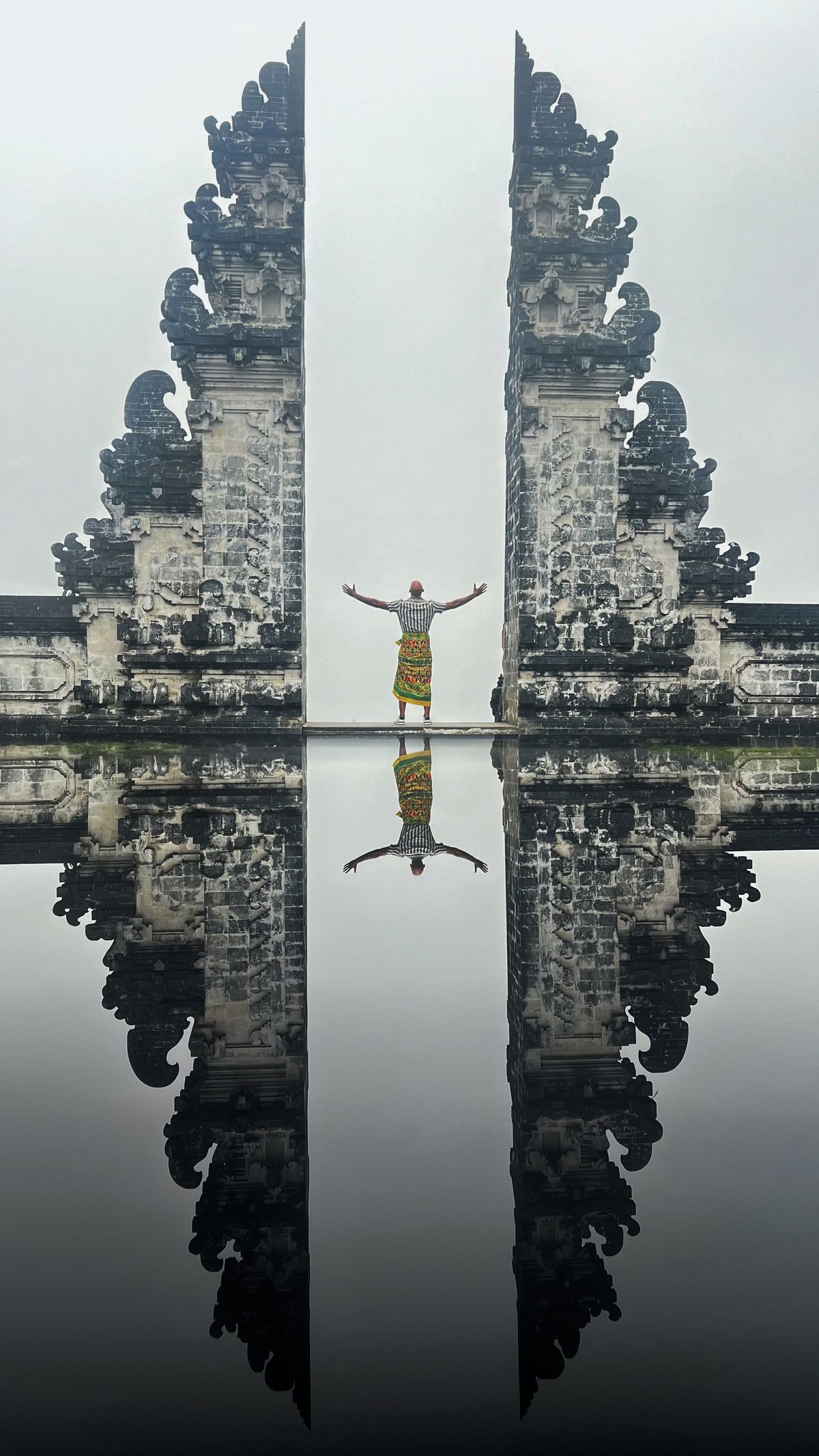 A person stands with arms out in the Lempuyang gate with a mirror reflection below and a plain white cloudy sky, no volcano visible