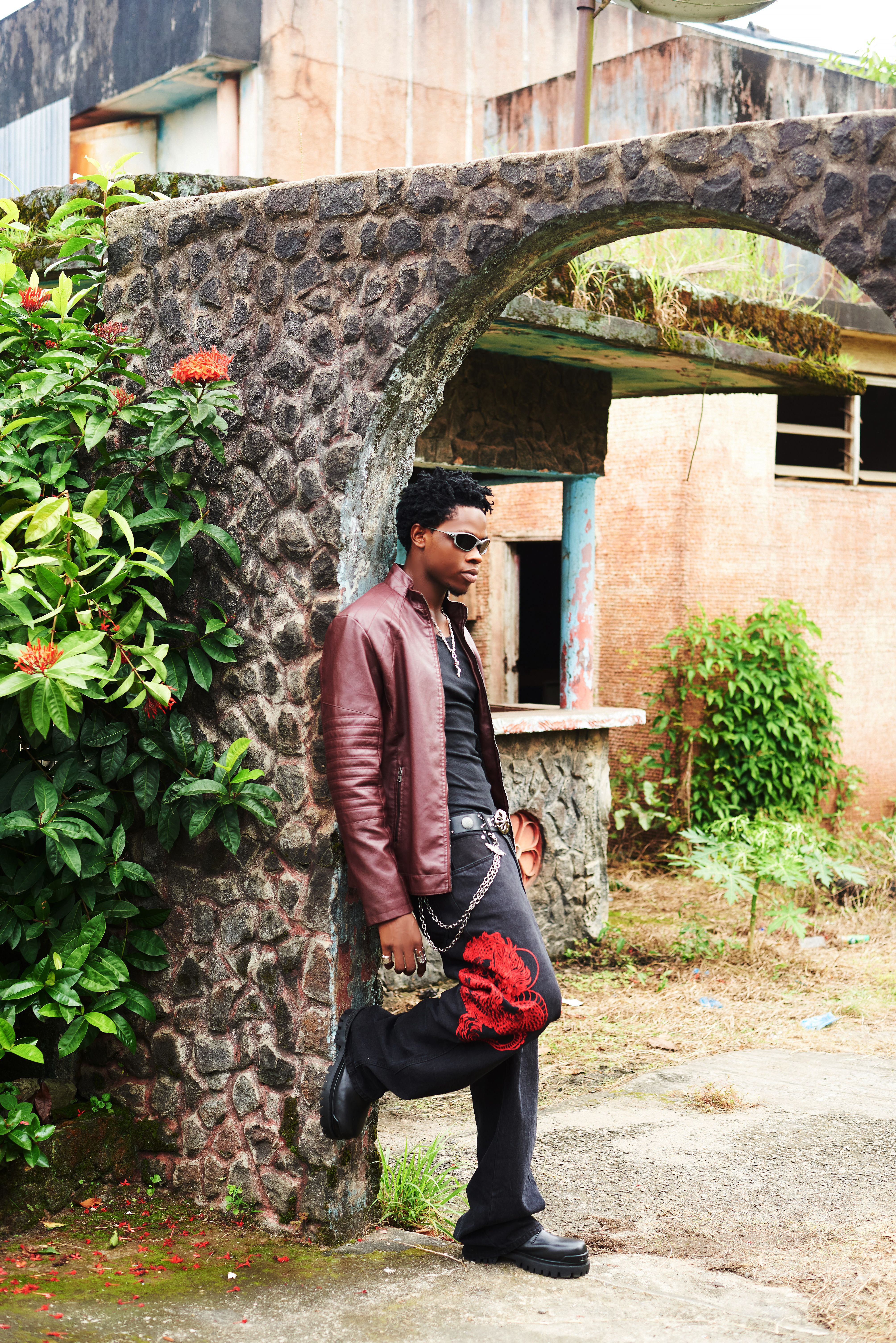 Man in fashion attire leaning against stone archway with lush greenery.