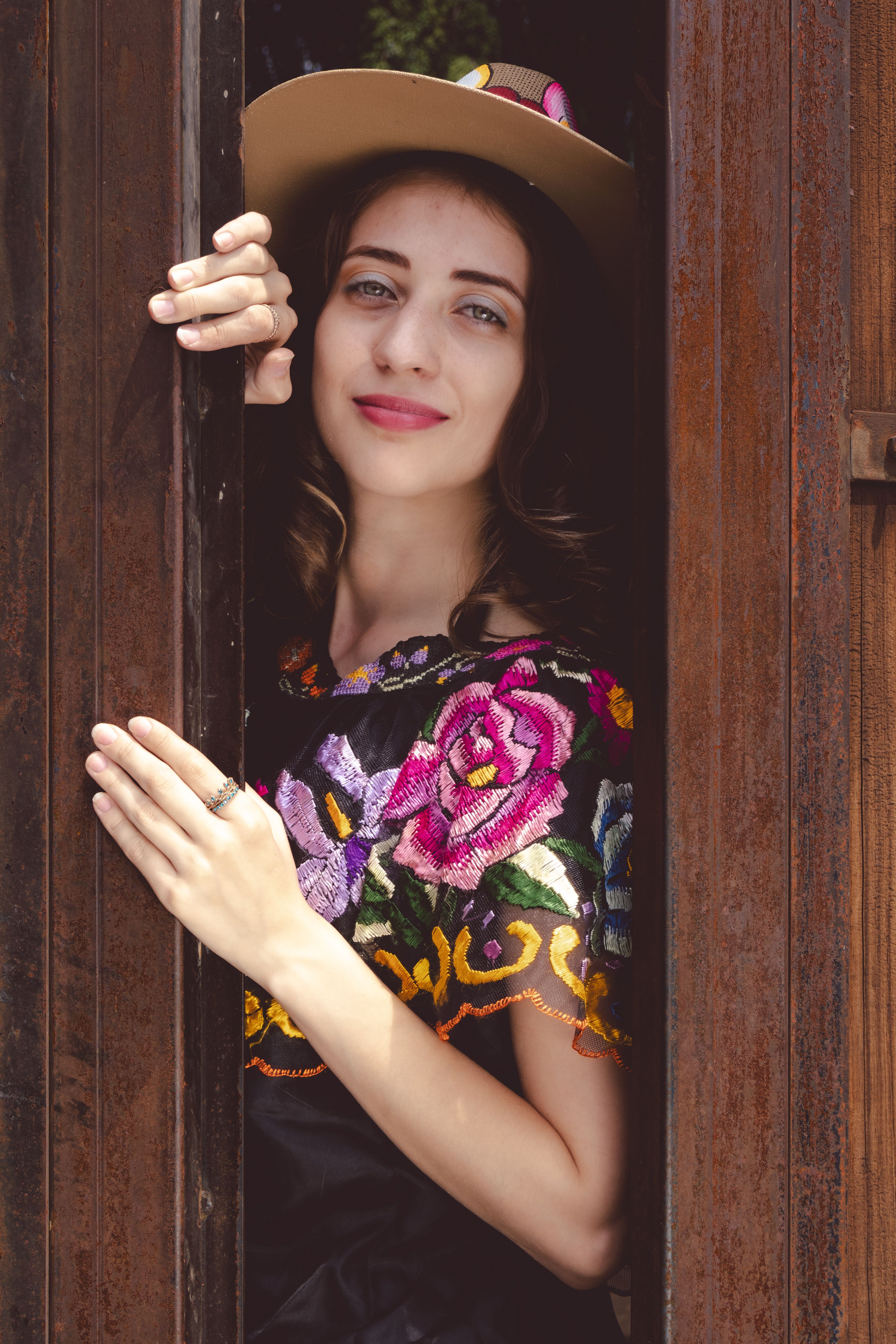 Smiling woman peeks from a wooden door, wearing a colorful embroidered top and hat.