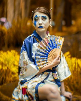 A woman in traditional costume and makeup holds a fan in an outdoor park setting.