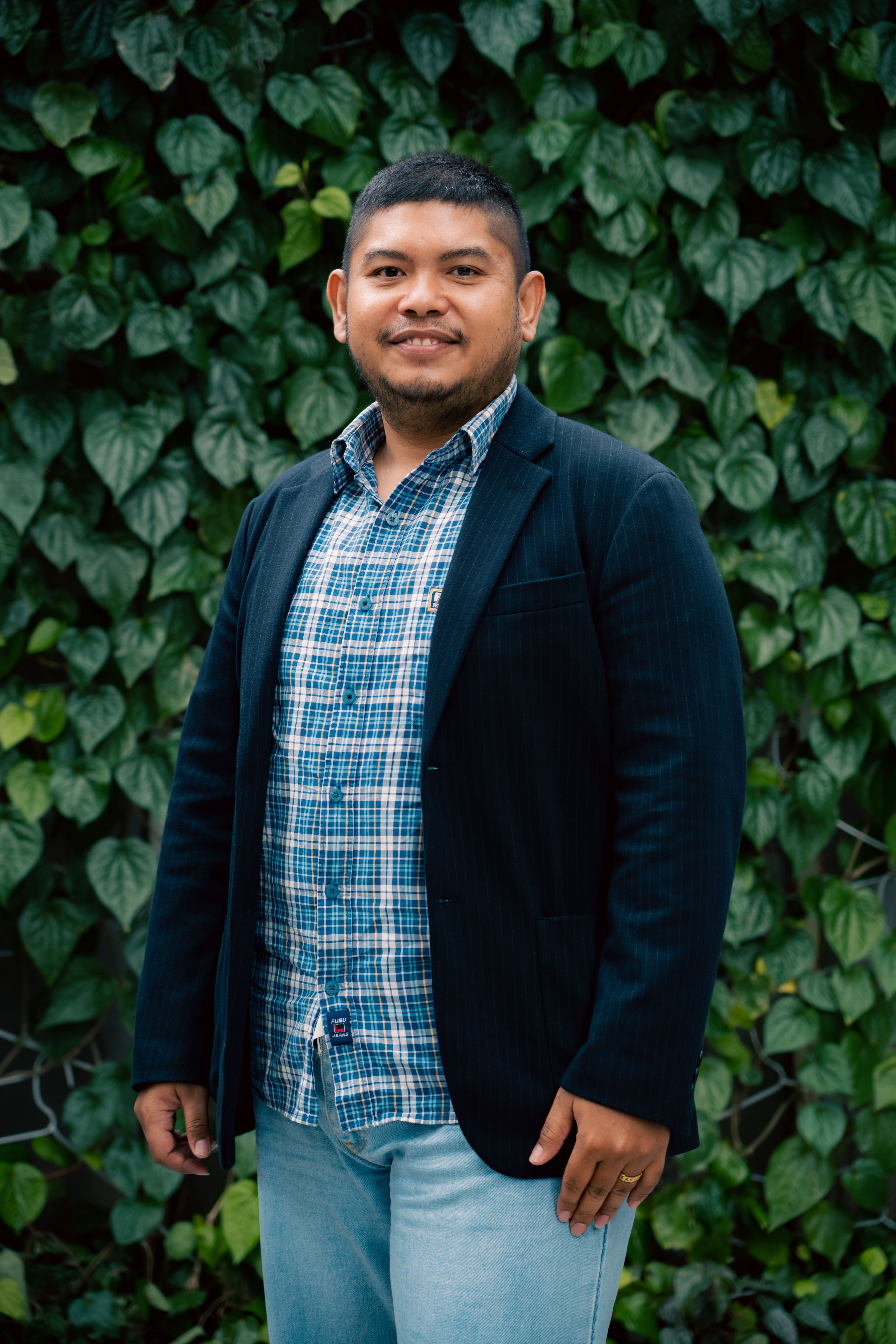 A man smiling in front of a lush green leafy background outdoors, wearing casual attire.