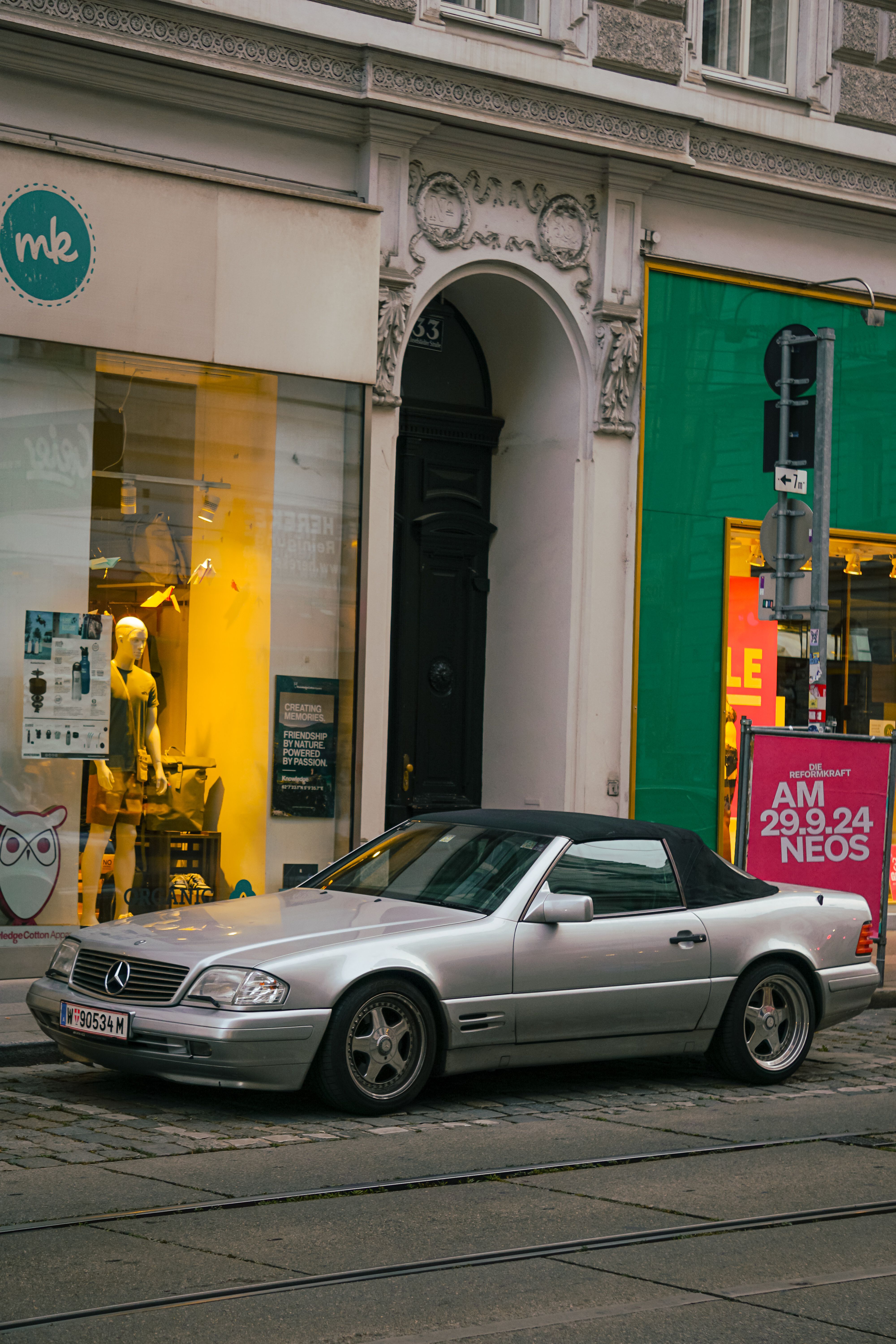Silver convertible car parked on a city street with vibrant storefronts and architecture.