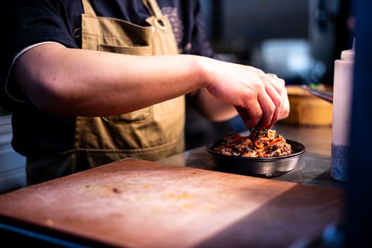 A close-up of a chef's hands garnishing a dish in an indoor kitchen setting, highlighting culinary artistry.