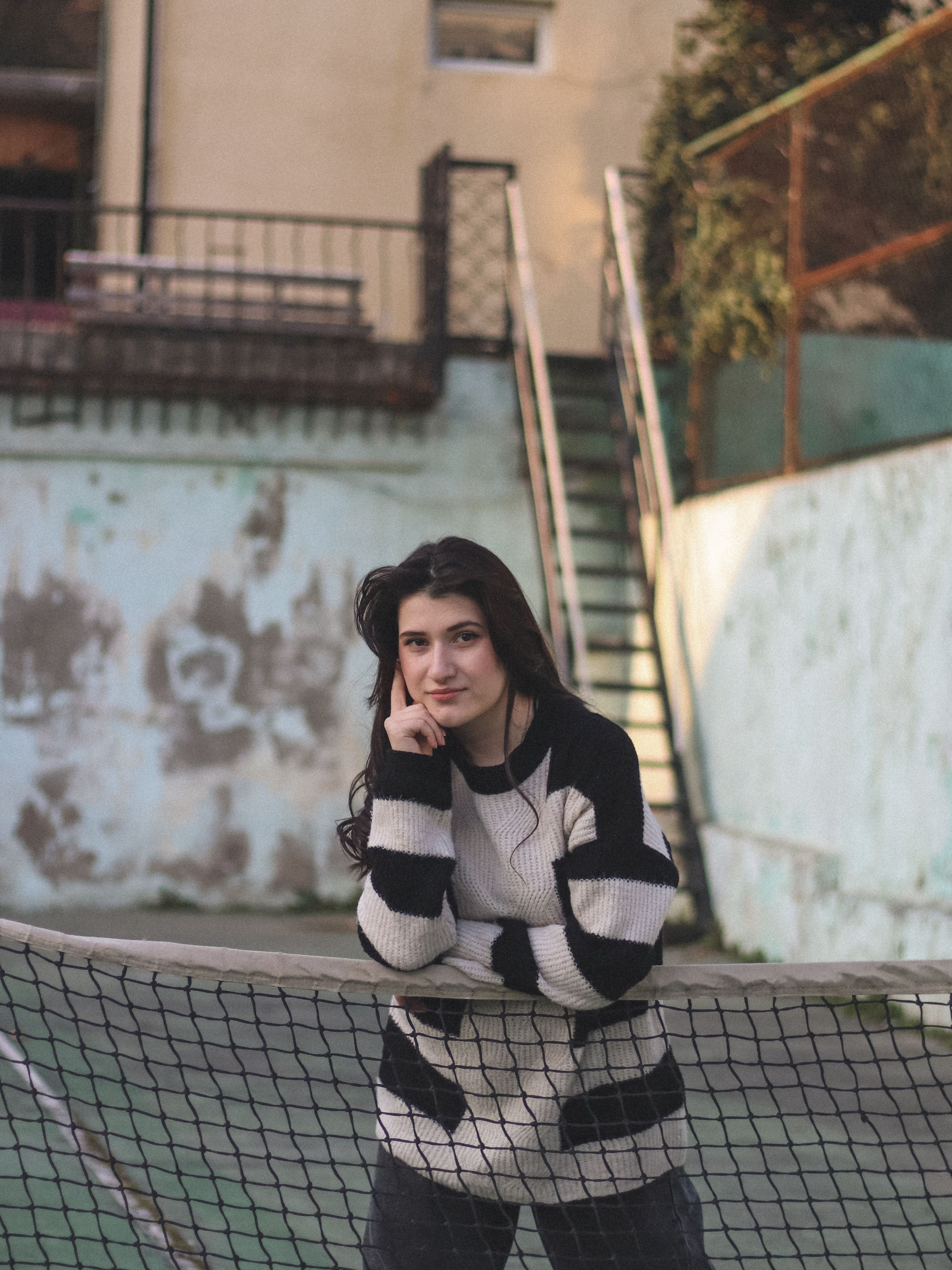 Portrait of a woman leaning on a tennis net wearing a striped sweater outdoors.