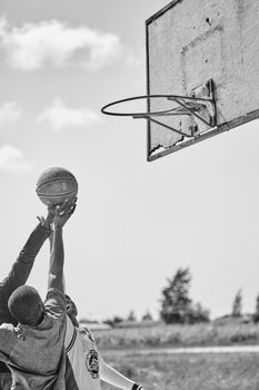 Two athletes compete for a basketball in an outdoor court in a dynamic black and white shot.