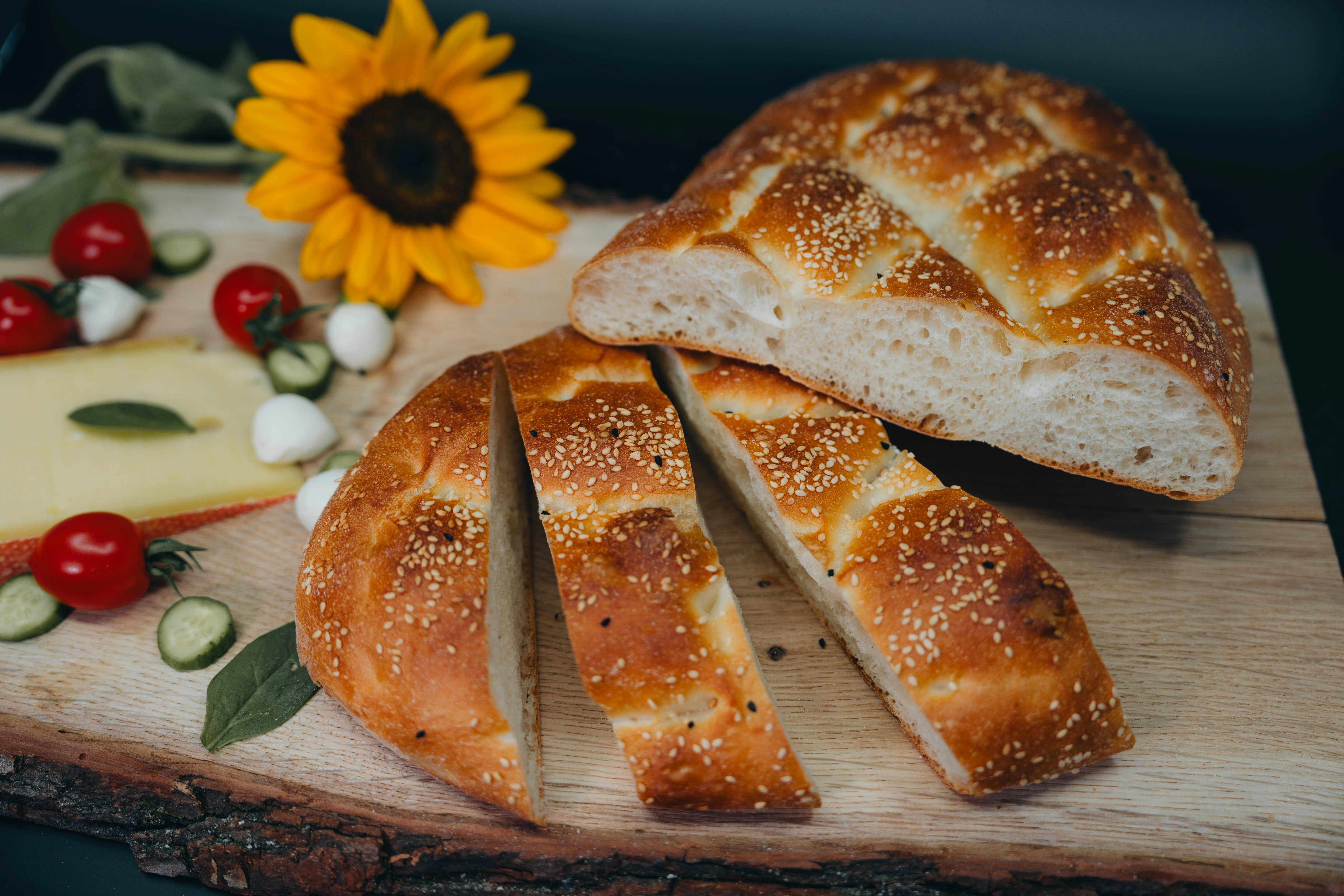 Freshly baked sesame bread slices on a wooden board, surrounded by cheese, cherry tomatoes, cucumbers, mozzarella, and a sunflower for a rustic food presentation.