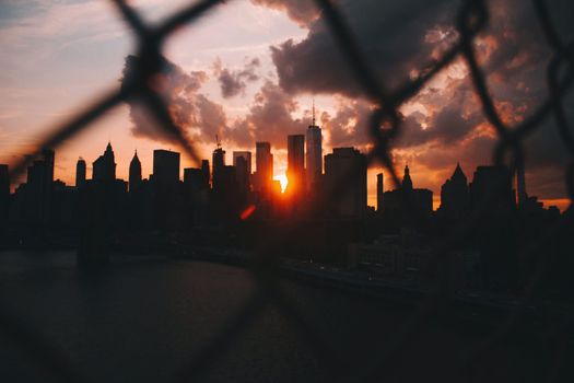 Dramatic sunset behind city skyline, viewed through a chain link fence with vibrant clouds.