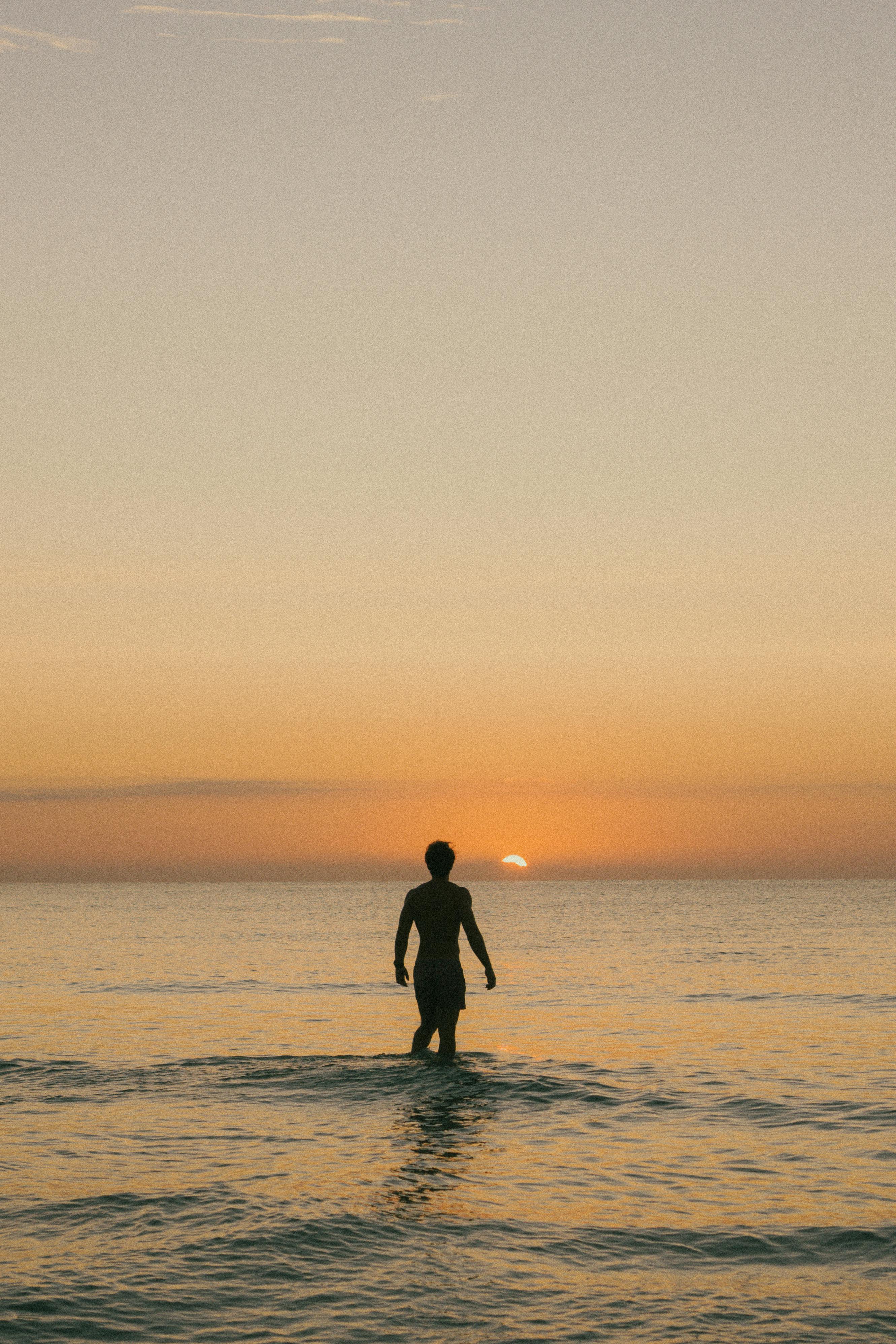A lone man wades into the calm sea during a warm sunset, creating a peaceful silhouette.