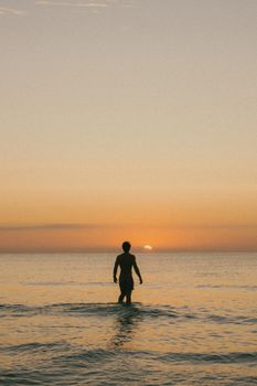 A lone man wades into the calm sea during a warm sunset, creating a peaceful silhouette.