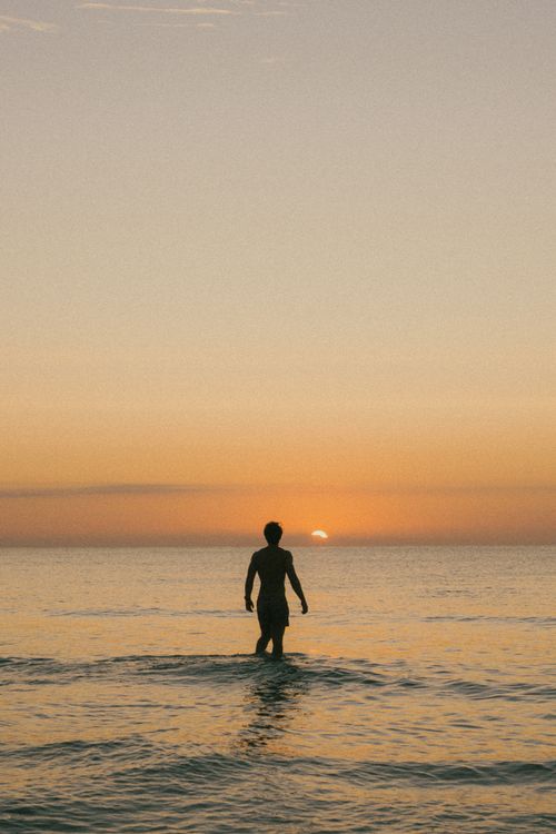 Free A lone man wades into the calm sea during a warm sunset, creating a peaceful silhouette. Stock Photo
