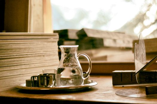 A peaceful still life scene with a glass water pitcher and books on a wooden table by the window.