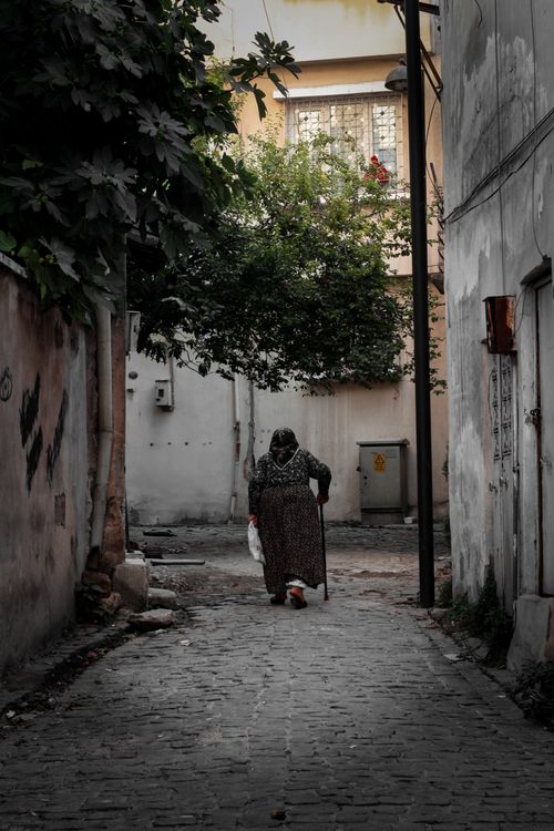 Free An elderly woman with a cane and groceries walks down a narrow alley between buildings. Stock Photo
