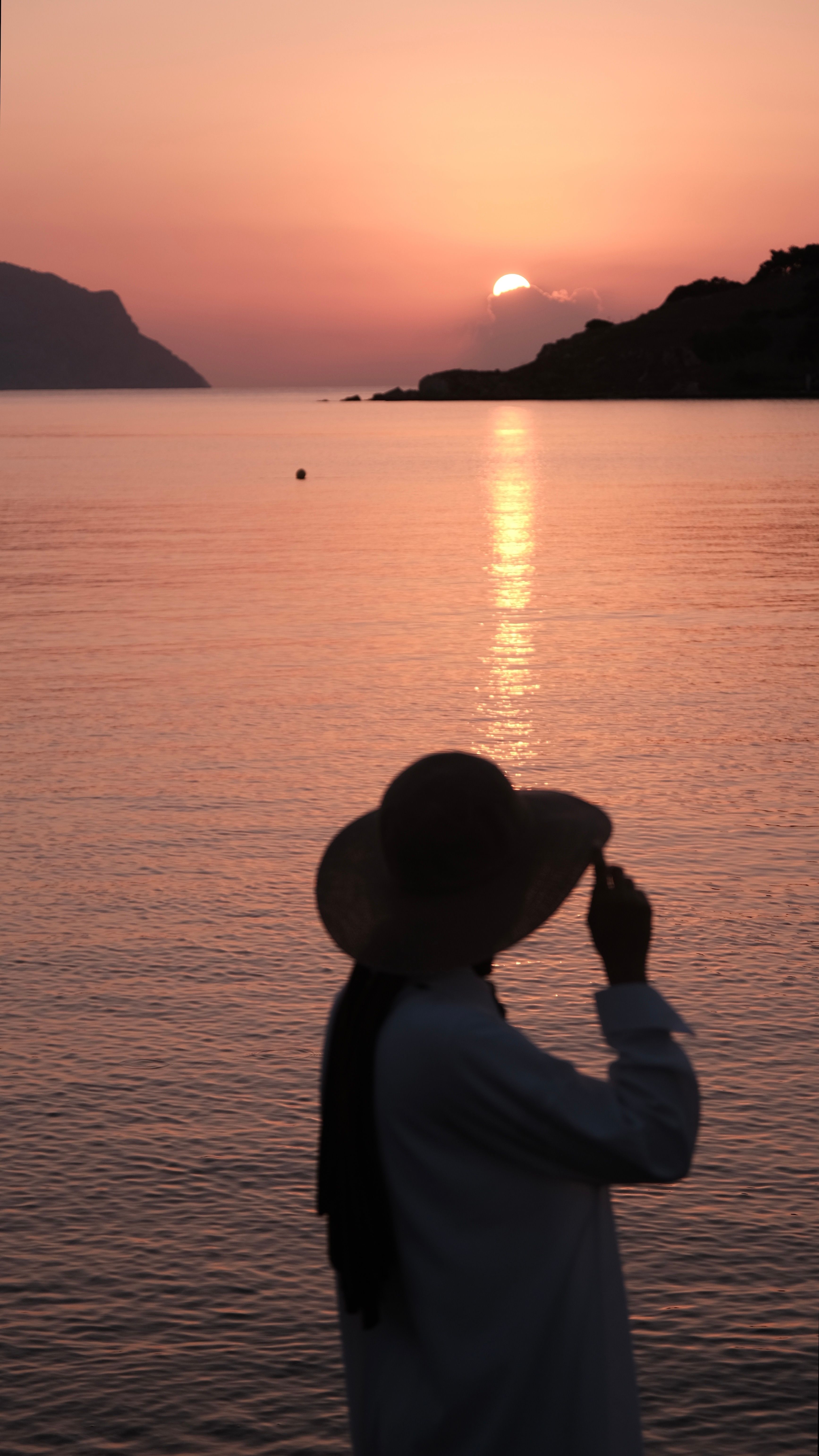 Silhouette of a person with hat at sunset, reflecting on calm sea.