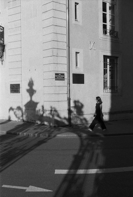 Black and white street photo of a woman walking in Nancy, France, captured with dramatic shadows.